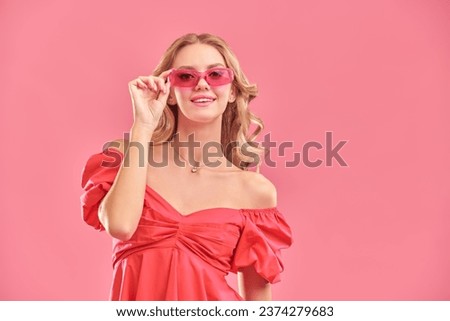 Young beauty. A cute attractive girl with blond hair poses in a pink dress and pink glasses and smiles. Pink studio background with copy space. Femininity, beauty and fashion.