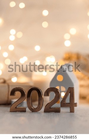 Happy new year 2024 postcard. Golden digits in front of Christmas lights, candles, gift box. White wooden background. Copy space for greeting text