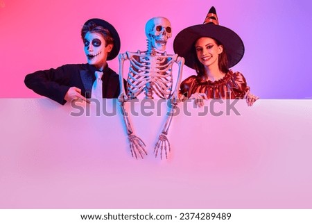 Pretty young witch and a sugar skull teenage boy are holding skeleton and a sign with copy space. Halloween celebration. Costumes, make-up and hairstyle. Studio portrait with mixed colorful lighting.
