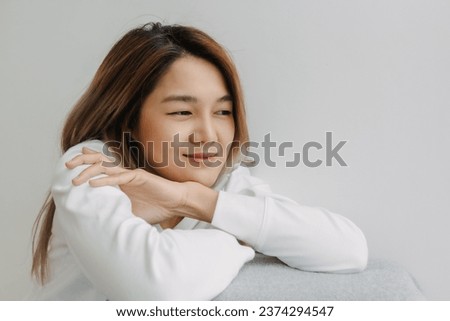 Asian Thai woman smiling face, thinking of good thing memories time, resting chin on arms at apartment room in winter alone. Royalty-Free Stock Photo #2374294547