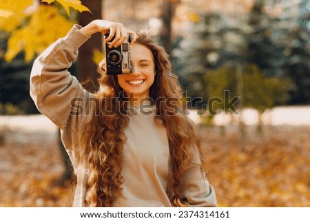Smiling young woman photographer with the photo camera shooting the forest trees with the yellow leaves at sunset