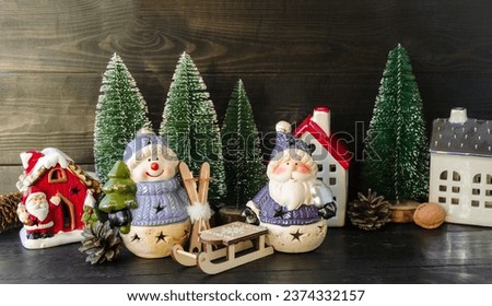 New Year card, porcelain toy snowman with a Christmas tree in his hands and Santa next to Santa's house and green fir trees and wooden sleigh.  Decorative decoration for home.  Background picture.