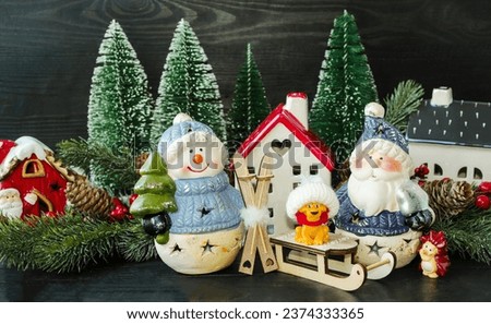 New Year card, porcelain toy snowman with a Christmas tree in his hands and Santa next to Santa's house and green fir trees and wooden sleigh.  Decorative decoration for home.  Background picture.
