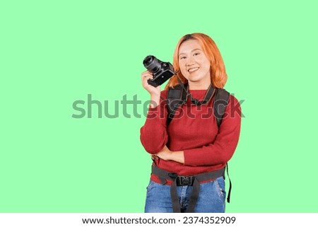 Young trendy photographer with red hair carrying camera backpack standing on isolated green background