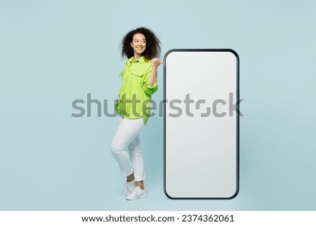 Full body young latin woman she wearing green shirt casual clothes point on big huge blank screen mobile cell phone smartphone with workspace area isolated on plain pastel light blue cyan background