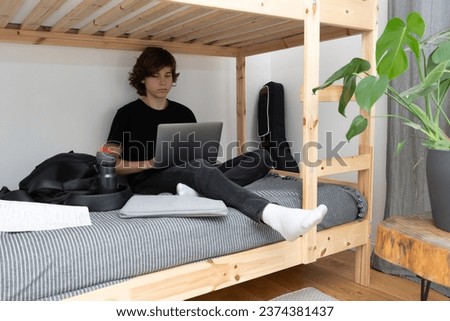 A teenage schoolboy or student is sitting on the lower tier of a bunk bed in his room with a laptop in his hands and doing homework. Royalty-Free Stock Photo #2374381437