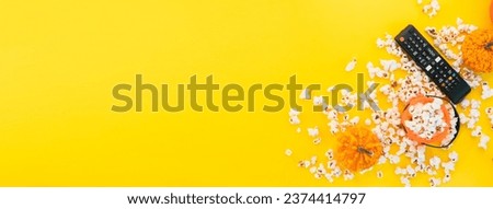 Remote control and red and white stripped box full of popcorn on a yellow background banner. Watching TV at home. Entertainment concept. Tv shows or movie night background with copy space.