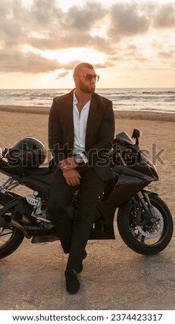 Elegantly dressed man in black suit leans on a sports motorcycle on a desolate beach against a cloudy sunset