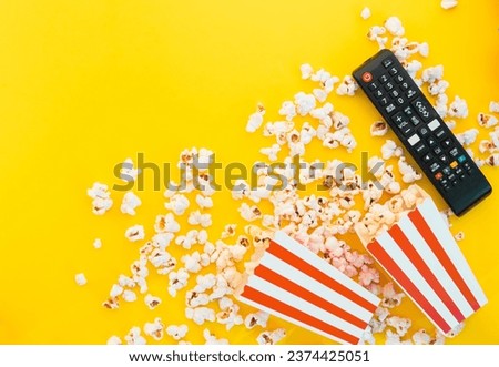 Remote control and red and white stripped box full of popcorn on a yellow background. Watching TV at home. Entertainment concept. Tv shows or movie night background with copy space.