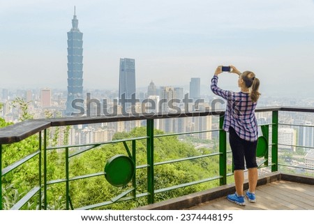 Young woman taking pictures and enjoying scenic view of Taipei from top of mountain. Awesome Taipei skyline, Taiwan. Wonderful cityscape. Skyscrapers and other modern buildings of downtown.