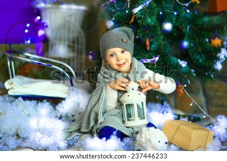 the girl in lilac jeans, in a knitted cap and with a knitted scarf sits on a sledge near a Christmas fir-tree with a toy and it is snowing