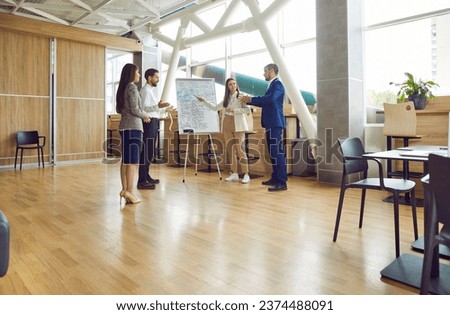 Professional business mentor and coach meeting with team of young people in modern office interior, standing in front of flip chart presentation board, giving master class, and highlighting key points Royalty-Free Stock Photo #2374488091
