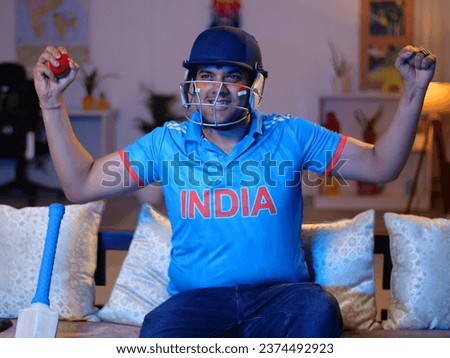 Excited young Indian man wearing cricket jersey and helmet - watching cricket match on tv, excitement. Indian man enthusiastically making supporting and winning gestures while watching a live crick...