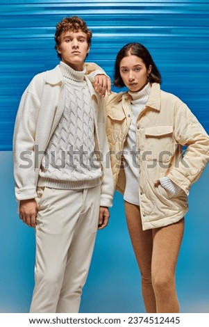 multiethnic couple in warm jackets and leather boots looking at camera on blue plastic backdrop
