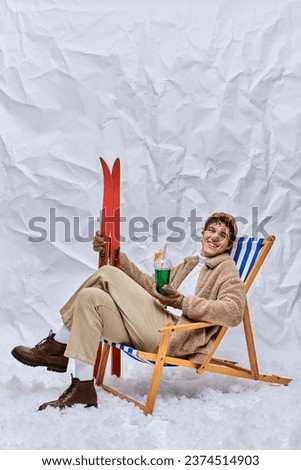 cheerful man in winter attire sitting in deck chair with apres-ski beverage and skis in snowy studio