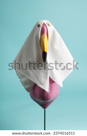 a plastic flamingo decoration disguised as a ghost with a sheet and false eyes on a turquoise background. Minimalist, trendy still life photography.