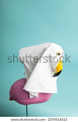 a plastic flamingo decoration disguised as a ghost with a sheet and false eyes on a turquoise background. Minimalist, trendy still life photography.