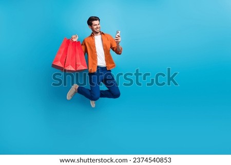Full body photo of handsome excited guy jumping use smart phone hold bags empty space isolated on blue color background
