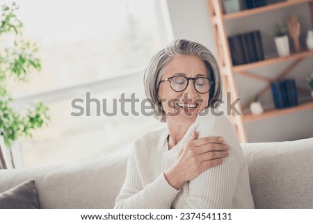 Photo of positive adorable lady pensioner sitting on comfy sofa hugging herself enjoying autumn cozy day home indoors