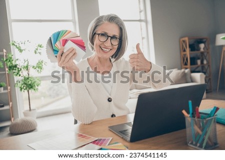 Photo of charming confident senior lady dressed white cardigan showing thumb up holding colors palette indoors house room