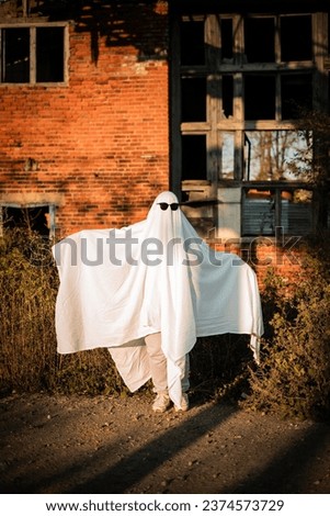 A man in a ghost costume made from a sheet and sunglasses stands near an abandoned building. Ghost Challenge 2021. Spooky season. Celebrating halloween.