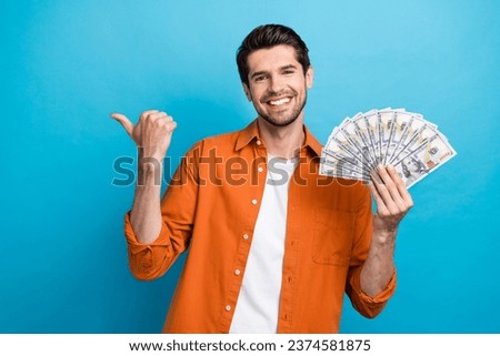Photo of cheerful excited man dressed orange shirt rising cash fan thumb empty space isolated blue color background
