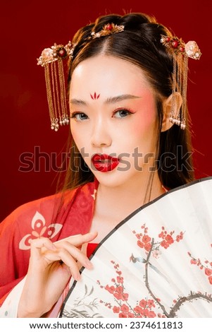 Beautiful young Asian woman with clean fresh skin wearing traditional cheongsam qipao dress holding fan posing on red background. Portrait of female model in studio. Happy Chinese new year.