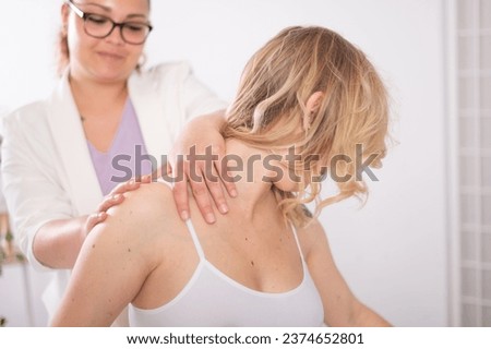 Crop smiling female therapist in glasses rubbing shoulder of blond haired client wearing white bra during physiotherapy session Royalty-Free Stock Photo #2374652801