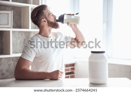 Handsome young bearded sportsman is drinking while preparing sport nutrition in kitchen at home Royalty-Free Stock Photo #2374664241