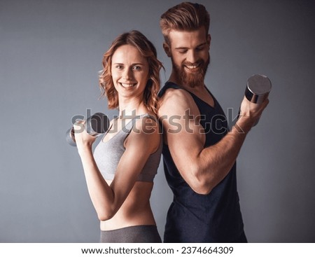 Beautiful young sports people are working out with dumbbells and smiling, on gray background