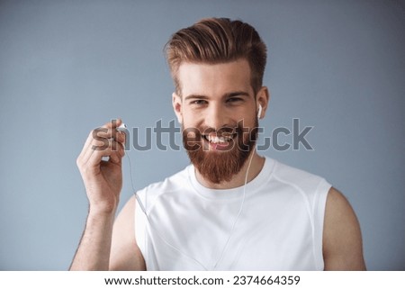 Portrait of handsome young bearded sportsman in earphones looking at camera and smiling, on gray background