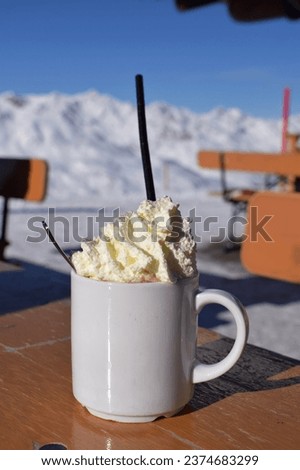 Delicious hot chocolate outdoors in snowy ski resort. Fluffy whipped cream in blank white mug with snowcapped mountain background. Ideal hot drink for a rest during skiing and snowboarding holiday