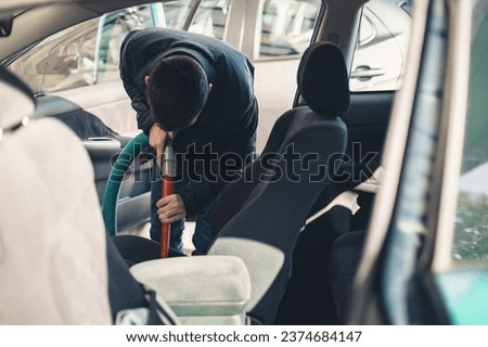 One young handsome caucasian brunette man vacuuming the front passenger seat inside the salon in his car at the city car wash on a clear spring sunny day, top view, close-up.