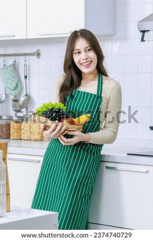 Asian young beautiful housewife in casual outfit with apron standing smiling holding mixed fresh organic fruits basket and vegetables basket in full decorated modern kitchen with ingredients at home.