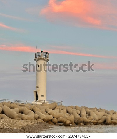 A view of the white lighthouse on the breakwater. The thin clouds behind the lighthouse are sunset, colored with pastel colors of light pink and orange medium tones. Royalty-Free Stock Photo #2374794037