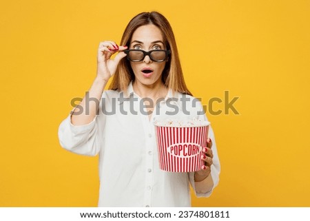 Young shocked astonished amazed stupefied pop-eyed woman lower 3d glasses wearing white casual clothes watching movie film hold bucket of popcorn in cinema isolated on plain yellow background studio