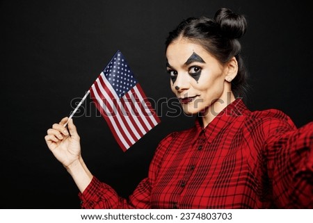 Young woman with Halloween makeup face art mask wear clown costume red dress do selfie shot mobile cell phone hold American flag isolated on plain black background studio. Scary holiday party concept