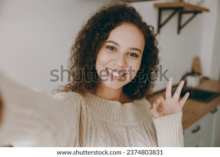 Close up young housewife woman wear casual clothes sweater doing selfie shot pov mobile cell phone show ok okay gesture stand near table in light kitchen at home alone. Lifestyle cooking food concept