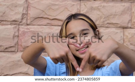 A cute preschool girl in a blue summer dress stands near a peach-colored brick wall with a smile on her face and makes a heart symbol from her hands, looking at the camera.