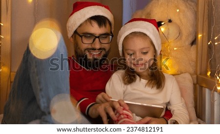 Happy family spend Christmas holiday time using digital tablet. Father and little girl in red santa hats sit in bed house and watch cartoons on tablet