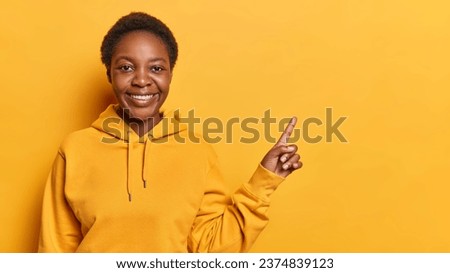 Indoor photo of young happy smiling broadly African american lady standing on left isolated on yellow background with blank space for your promotion wearing hoodie looking straight at camera Royalty-Free Stock Photo #2374839123