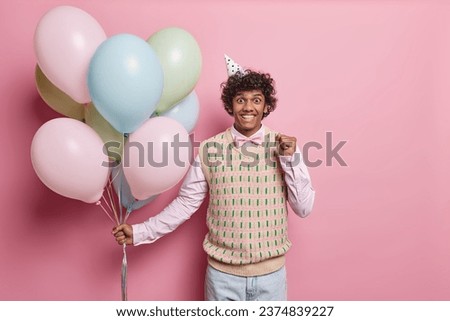 Cheerful young Hindu man with curly hair dressed in festive clothing clenches fist and celebrates special occasion holds bunch of inflated balloons comes on party isolated over pink background. Royalty-Free Stock Photo #2374839227