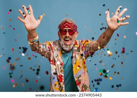Happy senior man in funky shirt and bandana throwing colorful confetti on blue background Royalty-Free Stock Photo #2374905443