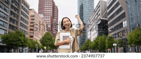 Portrait of happy asian woman stands with tablet near street road, cheering, raising hand up in triumph, celebrating.