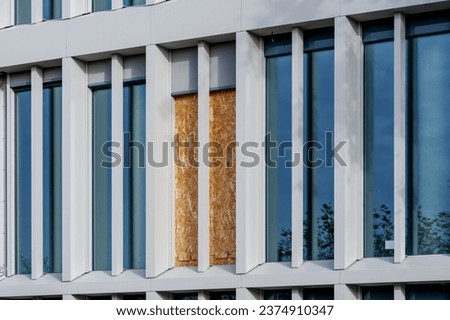 Outdoor exterior front view of modern facade with typical rectangular modern narrow vertical windows and one part of broken window which closed by OSB board. Royalty-Free Stock Photo #2374910347