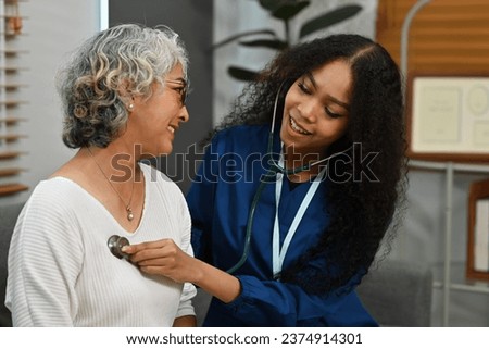 An African-Asian doctor wearing a scrub uniform listening to an older woman lung or heart sounds with a stethoscope during a visit at home, medical checkup and health care concept. Royalty-Free Stock Photo #2374914301