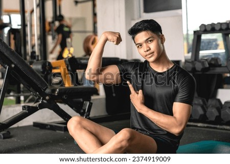 A young handsome and confident asian man flexing his bicep while sitting on the mat at the gym. Royalty-Free Stock Photo #2374919009
