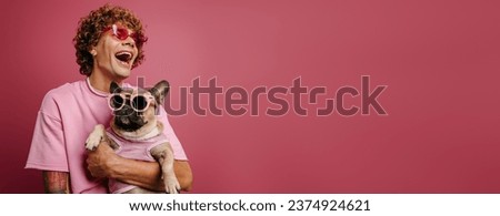 Joyful young curly hair man holding French Bulldog pet in funky glasses on pink background