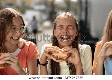 Portrait of joyful smiling teenage girls (14-15 years old) sitting at a cafe outdoors, enjoying hamburgers. Young ladies holding burgers, ready to eat them with pleasure.  Royalty-Free Stock Photo #2374967339