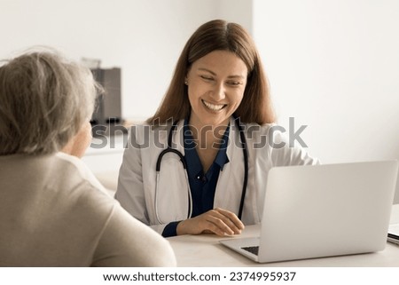 Smiling young doctor in uniform sit at desk talk to female retied client at medical checkup, show test result, explain treatment disease. Caring therapist use laptop to review patient health history Royalty-Free Stock Photo #2374995937